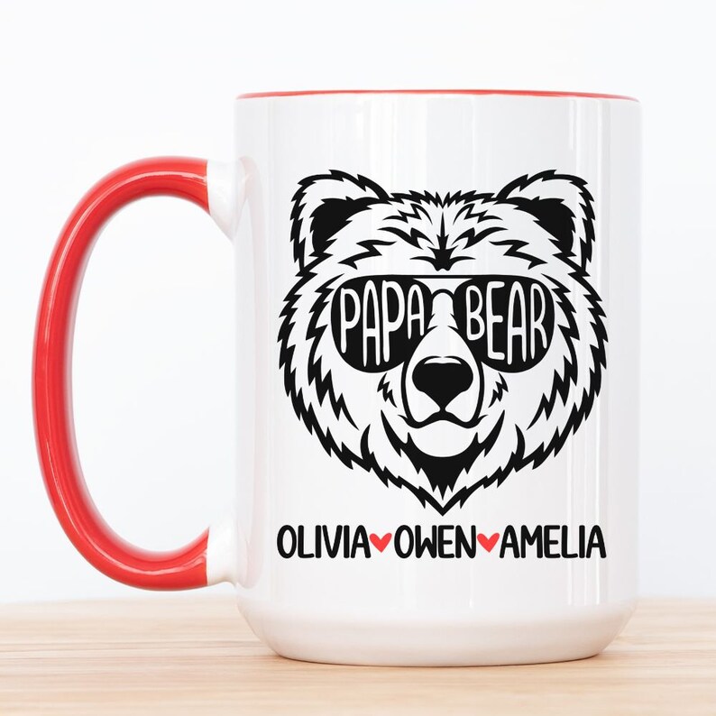 Op de afbeelding: Een witte keramische mok met een rode handgreep. De mok heeft een zwart-witte illustratie van een beer met een zonnebril met de tekst "Papa Bear" boven het hoofd van de beer. Onder de beer staat de tekst "Olivia Owen Amelia" in zwart met rode hartjes tussen de namen.
