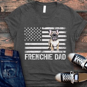 May include: Dark grey heather t-shirt with a white and grey American flag design. A French bulldog puppy is standing in the center of the flag. The text "FRENCHIE DAD" is printed below the flag.