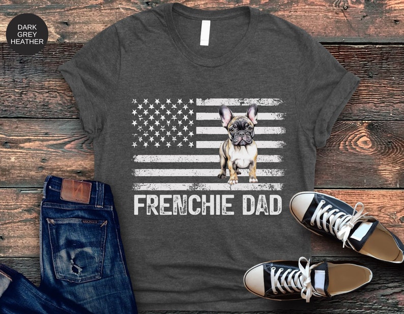 May include: Dark grey heather t-shirt with a white and grey American flag design. A French bulldog puppy is standing in the center of the flag. The text "FRENCHIE DAD" is printed below the flag.
