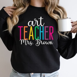 K&ouml;nnte beinhalten: Schwarzer Pullover mit dem Text "art teacher Mrs. Brown" in Regenbogenfarben.