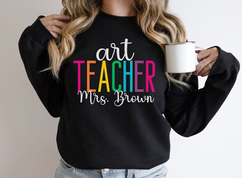 K&ouml;nnte beinhalten: Schwarzer Pullover mit dem Text "art teacher Mrs. Brown" in Regenbogenfarben.