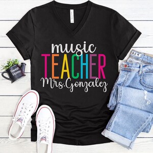 K&ouml;nnte beinhalten: Schwarzes V-Ausschnitt-T-Shirt mit dem Text "music teacher Mrs. Gonzalez" in Regenbogenfarben. Der Text ist in einer fetten, verspielten Schriftart.