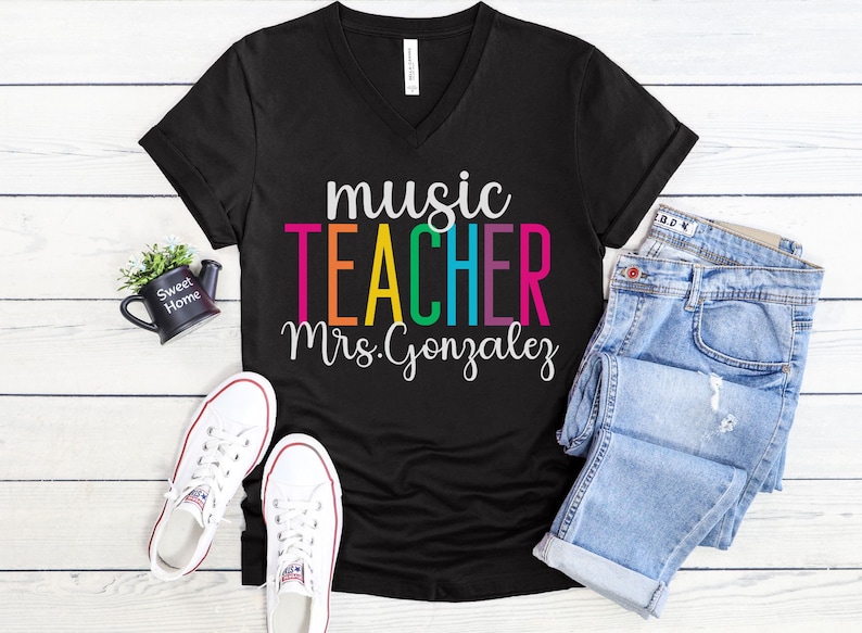 K&ouml;nnte beinhalten: Schwarzes V-Ausschnitt-T-Shirt mit dem Text "music teacher Mrs. Gonzalez" in Regenbogenfarben. Der Text ist in einer fetten, verspielten Schriftart.