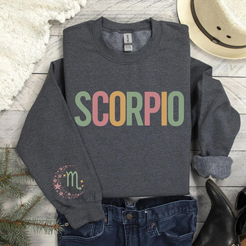 May include: A dark gray sweatshirt with the word "SCORPIO" printed in rainbow colors across the chest. The left sleeve has a small, colorful design of a crescent moon with a star and the letter "m" inside.