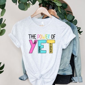 May include: Pale grey t-shirt with the text "THE POWER OF YET" in colourful block letters. The word "YET" is in pink, blue, and yellow. The shirt is hanging on a wooden hanger.