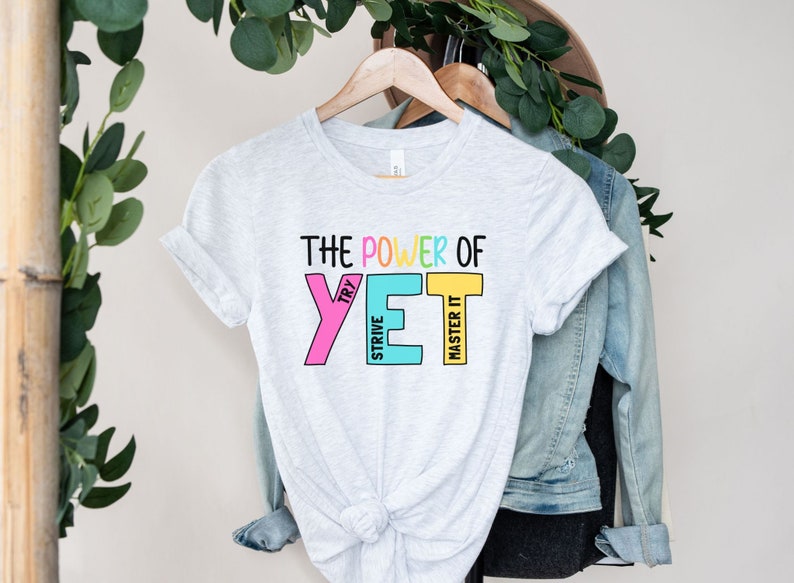 May include: Pale grey t-shirt with the text "THE POWER OF YET" in colourful block letters. The word "YET" is in pink, blue, and yellow. The shirt is hanging on a wooden hanger.