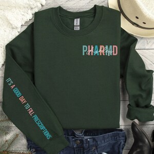 May include: A dark green sweatshirt with the text "PHARM" in colorful letters and the name "Ashley" written in cursive underneath. The left sleeve has the text "IT'S A GOOD DAY TO FILL PRESCRIPTIONS" in colorful letters.