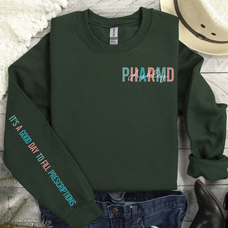 May include: A dark green sweatshirt with the text "PHARM" in colorful letters and the name "Ashley" written in cursive underneath. The left sleeve has the text "IT'S A GOOD DAY TO FILL PRESCRIPTIONS" in colorful letters.
