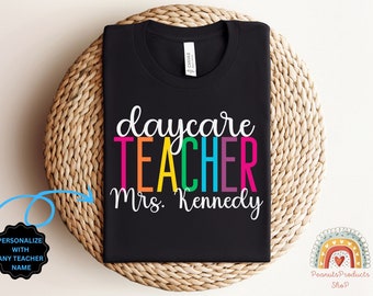 May include: Black t-shirt with the text "daycare teacher" in rainbow colors and "Mrs. Kennedy" in white below. The shirt is on a woven basket.
