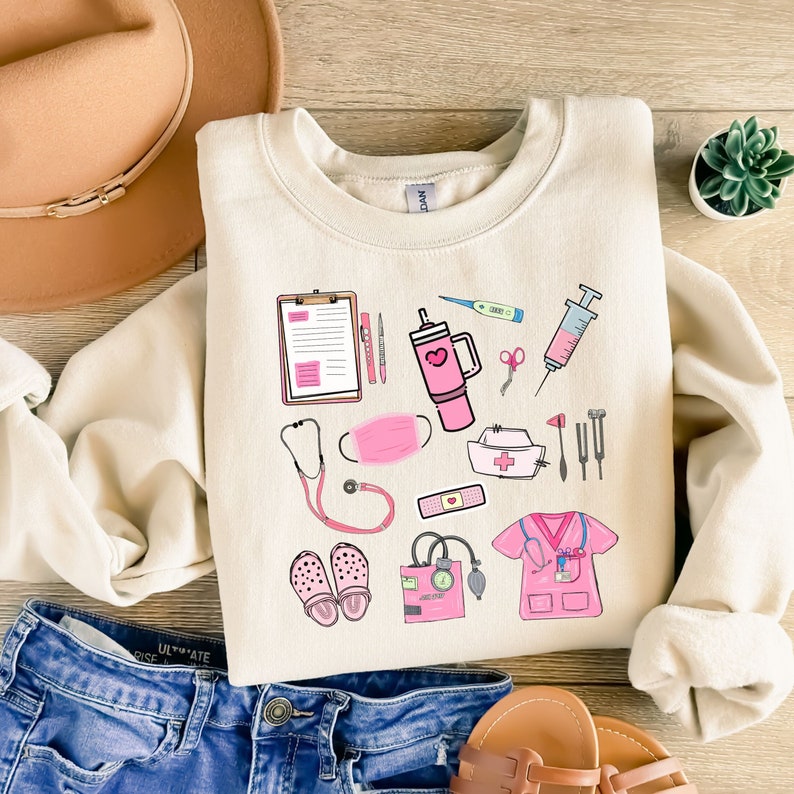 K&ouml;nnte beinhalten: Cremefarbenes Sweatshirt mit rosa medizinischen Illustrationen, darunter ein Stethoskop, eine Gesichtsmaske und medizinische Werkzeuge. Das Sweatshirt wird mit einer blauen Jeans und einem braunen Hut pr&auml;sentiert. Die Illustrationen sind im Cartoon-Stil.