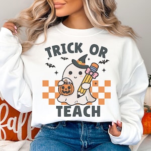 May include: White sweatshirt with a graphic of a ghost wearing a witch hat and holding a pencil and a pumpkin. The text "Trick or Teach" is printed on the sweatshirt.