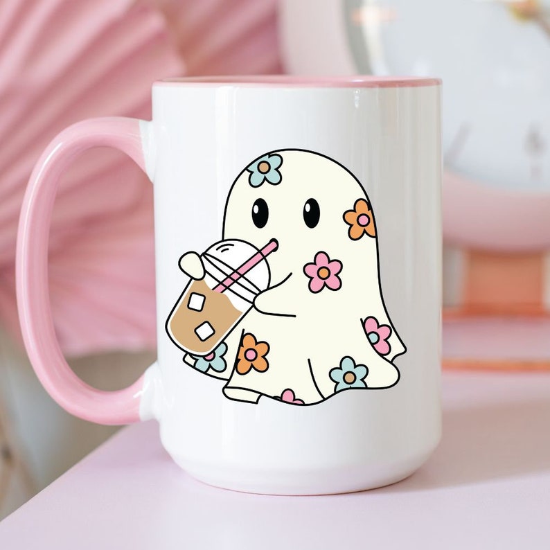 Op de afbeelding: Een witte keramische mok met een roze handvat. De mok heeft een cartoon-geest met bloemenpatronen die een kopje ijskoffie vasthoudt.