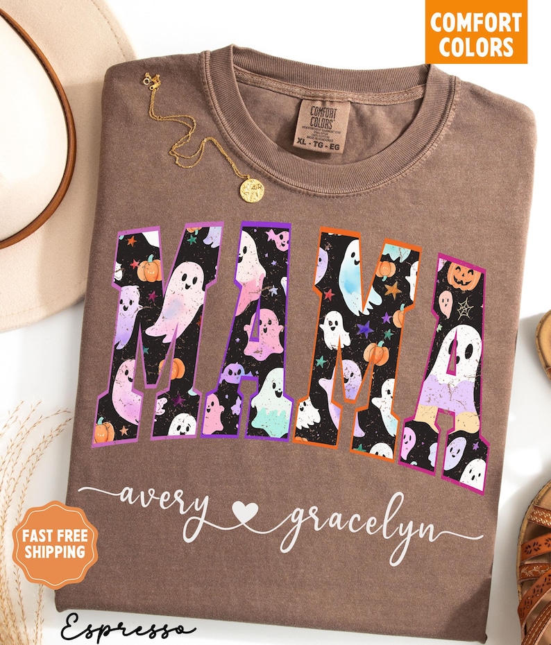 May include: Espresso-colored t-shirt with the word "MAMA" in a Halloween-themed design. The letters are filled with ghosts, pumpkins, and stars. The names "avery" and "gracelyn" are written below the word "MAMA".