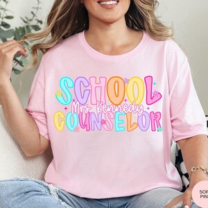 May include: A light pink t-shirt with the words "SCHOOL Mrs. Kennedy COUNSELOR" in colorful, bubble letters. The design includes flowers and stars. The shirt is a soft pink color.