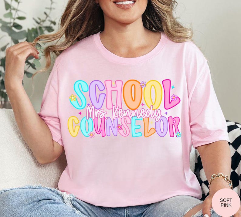 May include: A light pink t-shirt with the words "SCHOOL Mrs. Kennedy COUNSELOR" in colorful, bubble letters. The design includes flowers and stars. The shirt is a soft pink color.