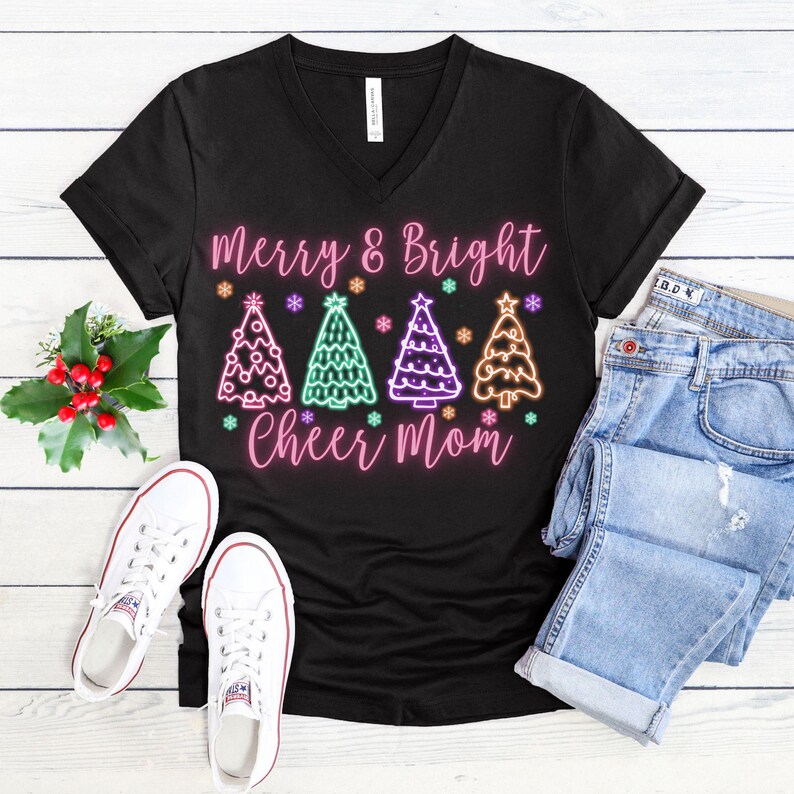 Pu&ograve; includere: Maglietta nera a collo a V con una grafica rosa di alberi di Natale e il testo "Merry & Bright Cheer Mom".