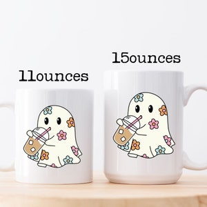 Op de afbeelding: Twee witte keramische mokken met een spookontwerp. Het spook draagt een wit laken met pastelkleurige bloemen en houdt een kopje ijskoffie vast. De mokken zijn gelabeld met "11 ounces" en "15 ounces".