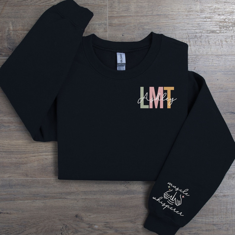 May include: Black sweatshirt with the text "LMT Ashley" on the front and "muscle whisperer" with a hand symbol on the sleeve.