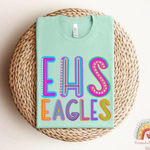 Puede incluir: Una camiseta verde claro con el texto "EHS Eagles" en letras de colores y contorneadas. Las letras est&aacute;n decoradas con peque&ntilde;os puntos y tienen un estilo retro y juguet&oacute;n.