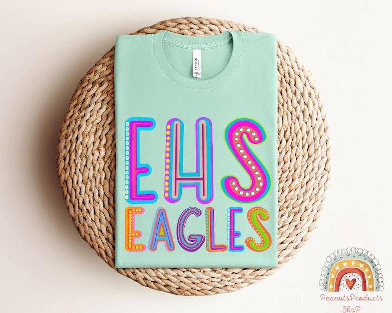 Puede incluir: Una camiseta verde claro con el texto "EHS Eagles" en letras de colores y contorneadas. Las letras est&aacute;n decoradas con peque&ntilde;os puntos y tienen un estilo retro y juguet&oacute;n.