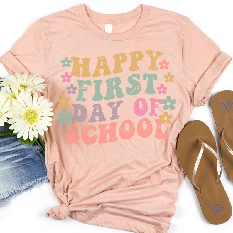 Puede incluir: Una camiseta rosa con el texto "Happy First Day of School" en una fuente retro con flores de colores que rodean el texto.