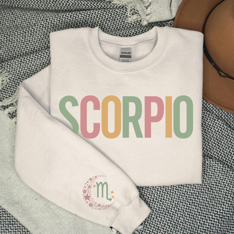 May include: A cream colored sweatshirt with the word "SCORPIO" printed in rainbow colors. The word is in a bold, blocky font. The sweatshirt has a small, embroidered design of the Scorpio zodiac symbol on the left sleeve.