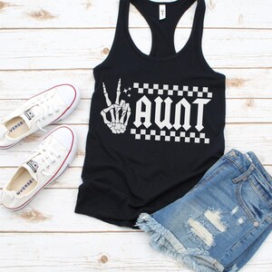 May include: Black tank top with a white skeleton hand making a peace sign and the word "AUNT" in a gothic font with a checkered pattern behind it.