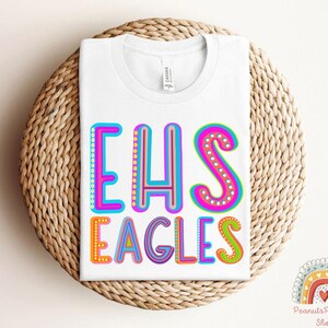Puede incluir: Camiseta blanca con el texto "EHS Eagles" en letras de colores y contorneadas. Las letras est&aacute;n decoradas con peque&ntilde;os puntos.