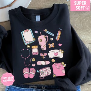 May include: Black sweatshirt with a colorful, floral-themed medical illustration. The design includes a stethoscope, pills, bandages, a nurse's cap, and a pink tumbler. The words "SUPER SOFT" are in a pink box.