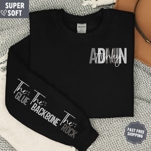 May include: Black sweatshirt with the text "ADMIN Ashley" in gray and white. The sleeve features the phrases "The Glue", "The Backbone", and "The Rock". The sweatshirt is labeled "Super Soft".