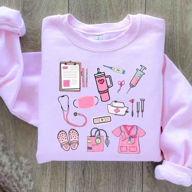 K&ouml;nnte beinhalten: Ein hellrosa Sweatshirt mit medizinischen Illustrationen in Rosa und Schwarz. Das Design umfasst ein Stethoskop, einen Klemmbrett, eine Gesichtsmaske, eine Krankenschwesterm&uuml;tze und andere medizinische Werkzeuge. Das Sweatshirt hat einen Rundhalsausschnitt und lange &Auml;rmel.