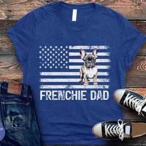 May include: A blue t-shirt with a distressed American flag design featuring a French Bulldog. The text "FRENCHIE DAD" is printed below the flag.