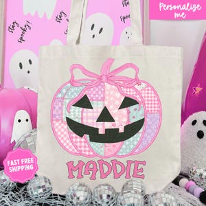 May include: A beige tote bag featuring a patchwork pumpkin design in pink, blue, and purple hues, with a black jack-o'-lantern face. The name "MADDIE" is printed below the pumpkin in pink letters. The bag has a pink bow at the top.