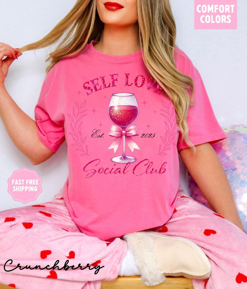 May include: Pink t-shirt with a graphic of a wine glass with a bow and the text "Self Love Social Club Est. 2025".