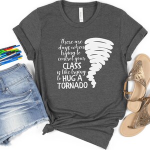 May include: A dark gray t-shirt with a white graphic of a tornado and the text "There are days when trying to control your CLASS is like trying to HUG A TORNADO".