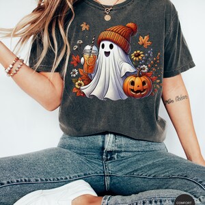 May include: A gray t-shirt with a white ghost wearing a brown and orange knit hat holding a glass of orange juice. The ghost is standing next to a jack-o-lantern with a happy face. The shirt has a fall-themed design with leaves, flowers, and berries.
