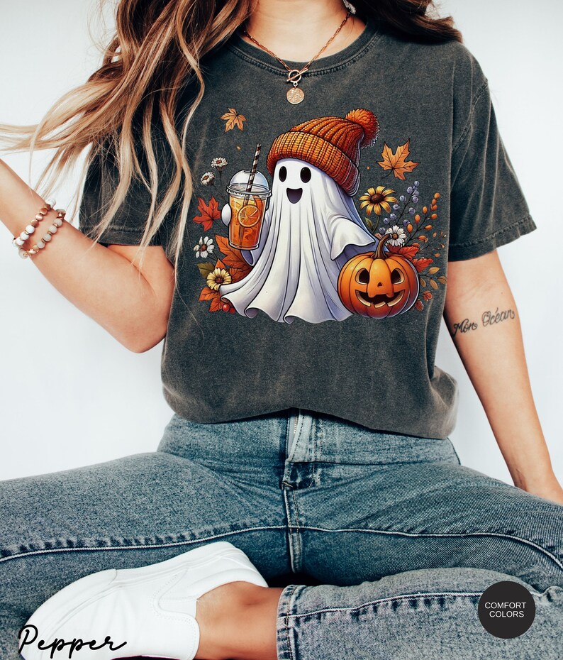 May include: A gray t-shirt with a white ghost wearing a brown and orange knit hat holding a glass of orange juice. The ghost is standing next to a jack-o-lantern with a happy face. The shirt has a fall-themed design with leaves, flowers, and berries.