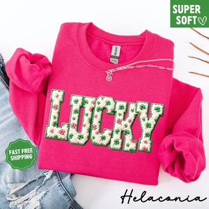 Pu&ograve; includere: Una felpa rosa acceso con la parola "LUCKY" ricamata sul davanti. Le lettere sono riempite con un motivo floreale di trifogli verdi e fiori rosa. Una collana d'argento &egrave; drappeggiata sopra. Le parole "SUPER SOFT" sono in una scatola verde.