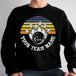 K&ouml;nnte beinhalten: Schwarzes Sweatshirt mit einer Retro-Grafik von Bowling-Pins und einer Bowlingkugel. Die Grafik enth&auml;lt einen Sonnenuntergang mit gelben, orangefarbenen und roten Streifen. Der Text "Your Team Name" ist um die Grafik herum geschrieben, mit dem Namen "Kevin" auf der Bowlingkugel.