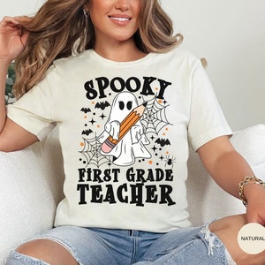 May include: White t-shirt with a black and orange graphic of a ghost holding a pencil with the text "Spooky First Grade Teacher".
