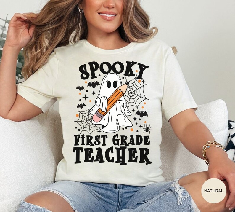 May include: White t-shirt with a black and orange graphic of a ghost holding a pencil with the text "Spooky First Grade Teacher".