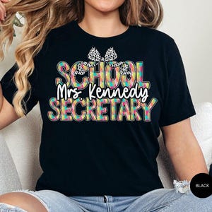 Może przedstawiać: Czarny T-shirt z kolorowym, wzorzystym nadrukiem, na kt&oacute;rym widnieje napis "School Mrs. Kennedy Secretary" z czarną i białą kokardką na g&oacute;rze.