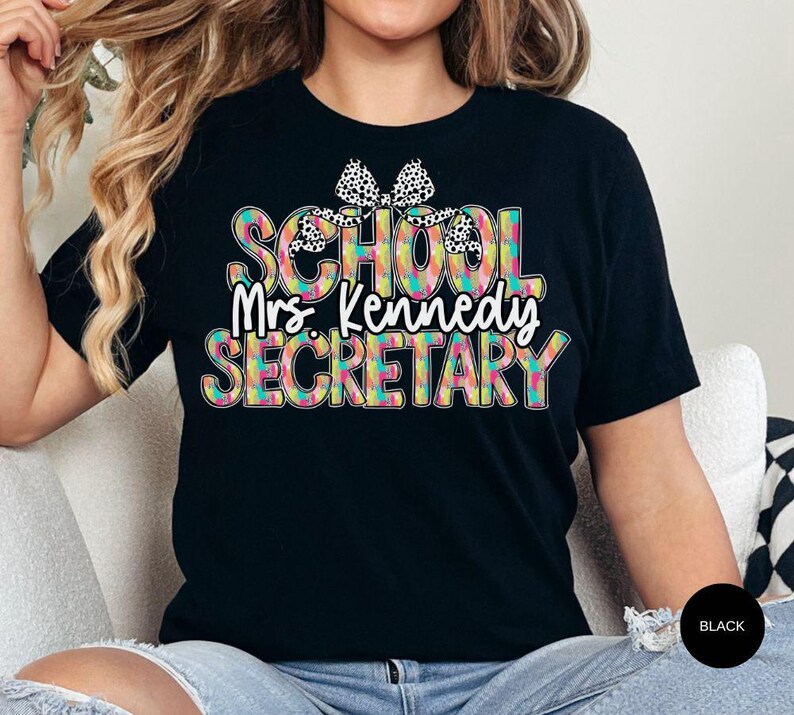 Może przedstawiać: Czarny T-shirt z kolorowym, wzorzystym nadrukiem, na kt&oacute;rym widnieje napis "School Mrs. Kennedy Secretary" z czarną i białą kokardką na g&oacute;rze.