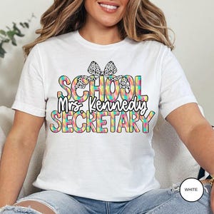 Może przedstawiać: Biała koszulka z kolorową grafiką, na kt&oacute;rej widnieje napis "School Mrs. Kennedy Secretary" z czarną i białą kokardką na słowie "School".