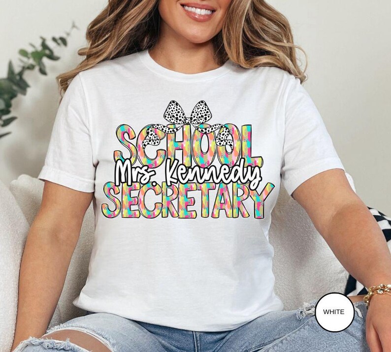 Może przedstawiać: Biała koszulka z kolorową grafiką, na kt&oacute;rej widnieje napis "School Mrs. Kennedy Secretary" z czarną i białą kokardką na słowie "School".