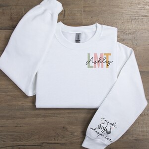 May include: White sweatshirt with the text "Ashley LMT" on the front and "muscle whisperer" with a hand illustration on the sleeve.