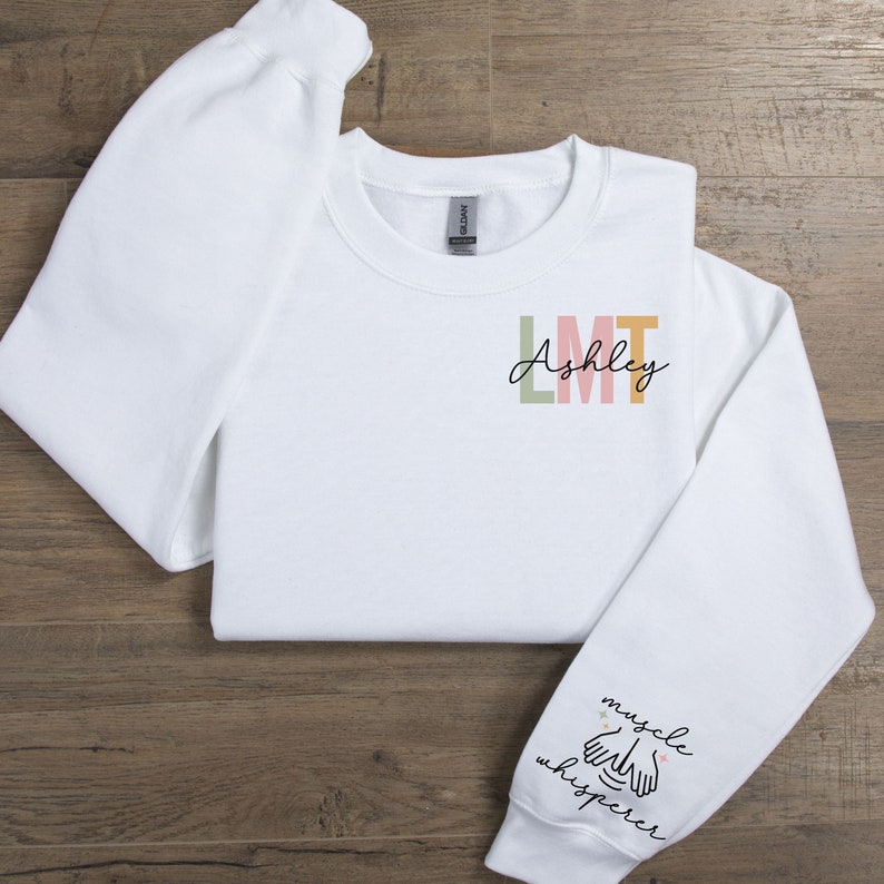May include: White sweatshirt with the text "Ashley LMT" on the front and "muscle whisperer" with a hand illustration on the sleeve.