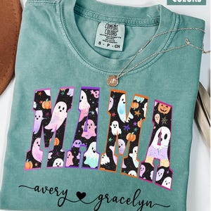 May include: Light green t-shirt with the word "MAMA" in a colorful Halloween-themed design featuring ghosts and pumpkins. The shirt also has the names "avery" and "gracelyn" in cursive script.