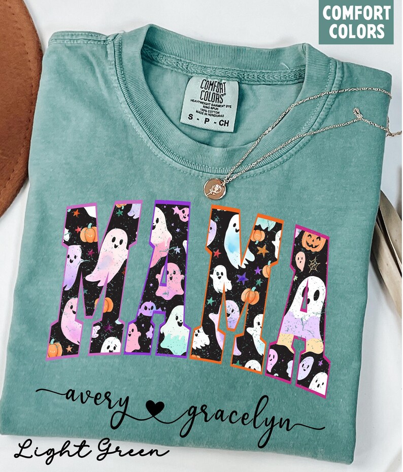May include: Light green t-shirt with the word "MAMA" in a colorful Halloween-themed design featuring ghosts and pumpkins. The shirt also has the names "avery" and "gracelyn" in cursive script.
