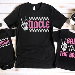 K&ouml;nnte beinhalten: Drei schwarze T-Shirts mit einem karierten Muster und einer Skeletthand. Die Shirts sagen "Onkel", "Cousin" und "Bad Two The Bone".